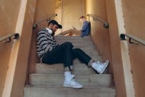 Vista laterale da vicino di due giovani caucasici alla moda seduti su gradini e che utilizzano smartphone in un magazzino abbandonato — Foto stock
