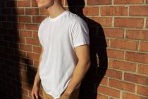 Vista lateral parte média de um jovem caucasiano vestindo uma camisa branca encostada a uma parede de tijolo ao sol — Fotografia de Stock