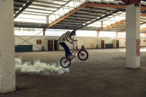 Side view of a young Caucasian man wearing a baseball cap riding and jumping on a BMX bike with a white smoke grenade attached to it, in an abandoned warehouse — Stock Photo