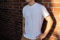 Vista lateral sección media de un joven hombre caucásico con una camiseta blanca de pie contra una pared de ladrillo con las manos en las caderas - foto de stock