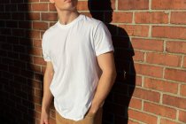 Side view mid section of a young Caucasian man wearing a white t shirt standing against a brick wall in the sun looking away — Stock Photo