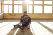 Frontansicht eines hippen jungen kaukasischen Mannes mit Baseballmütze und Kopfhörern, der auf dem Boden sitzt, an eine Säule in einer verlassenen Lagerhalle in der Sonne gelehnt, ein Smartphone in der Hand und Musik hörend, ein Skateboard neben sich — Stockfoto