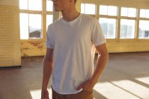 Vista frontal sección media de un joven caucásico vestido con una camiseta blanca de pie en un almacén abandonado al sol, mirando hacia otro lado, con la mano en el bolsillo - foto de stock