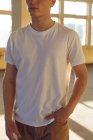Vista frontale metà sezione di un giovane caucasico che indossa una maglietta bianca in piedi in un magazzino abbandonato al sole, distogliendo lo sguardo, con la mano in tasca — Foto stock