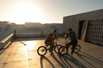 Vue latérale surélevée de deux jeunes hommes caucasiens assis sur des vélos BMX parlant sur le toit d'un entrepôt abandonné, rétroéclairé par le soleil couchant — Photo de stock
