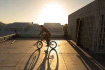 Вид сбоку на двух молодых кавказских мужчин, катающихся на велосипедах BMX на крыше заброшенного склада, подсвеченного заходящим солнцем — стоковое фото