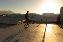 Side view of two young Caucasian men riding BMX bikes and doing tricks on the rooftop of an abandoned warehouse, backlit by the setting sun — Stock Photo