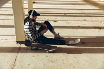 Seitenansicht eines hippen jungen kaukasischen Mannes mit Baseballmütze und Kopfhörern, der auf dem Boden sitzt, an eine Säule in einer verlassenen Lagerhalle in der Sonne gelehnt, ein Smartphone in der Hand und Musik hörend, ein Skateboard neben sich — Stockfoto
