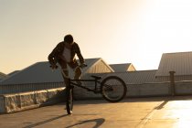 Vista lateral de un joven caucásico montando una bicicleta BMX y haciendo trucos en la azotea de un almacén abandonado, retroiluminado por el sol poniente - foto de stock