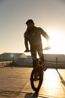 Vista frontal de cerca de un joven caucásico con una gorra de béisbol montada en una bicicleta BMX y haciendo trucos en la azotea de un almacén abandonado, retroiluminado por el sol poniente - foto de stock