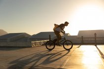 Vista lateral de um jovem caucasiano andando de bicicleta BMX no telhado de um armazém abandonado, iluminado pelo pôr-do-sol — Fotografia de Stock