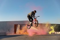 Vue latérale d'un jeune homme caucasien sautant dans les airs sur un vélo BMX faisant des tours sur le toit d'un entrepôt abandonné, avec des grenades fumigènes roses et jaunes en arrière-plan — Photo de stock