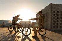 Vista lateral de dos jóvenes caucásicos sentados en bicicletas BMX hablando en la azotea de un almacén abandonado, retroiluminado por el sol poniente, con edificios al fondo - foto de stock