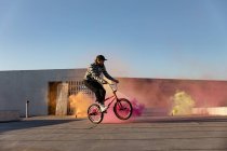 Vista lateral de um jovem caucasiano andando de bicicleta BMX fazendo truques no telhado de um armazém abandonado, com granadas de fumaça laranja, rosa e amarela no fundo — Fotografia de Stock
