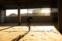 Vista frontal de um jovem caucasiano balanceando na roda dianteira de uma bicicleta BMX enquanto pratica truques em um armazém abandonado, retroiluminado pela luz solar — Fotografia de Stock