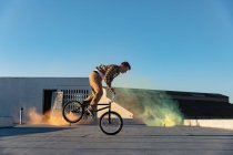 Vue latérale d'un jeune homme caucasien se balançant sur la roue avant d'un vélo BMX faisant des tours sur le toit d'un entrepôt abandonné, avec des grenades fumigènes vertes et orange en arrière-plan — Photo de stock