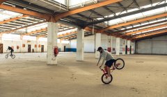 Side view of two young Caucasian men riding BMX bikes while practicing tricks in an abandoned warehouse, the rider in the foreground is balancing on the front wheel of his bike — Stock Photo
