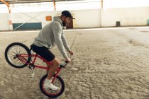 Nahaufnahme eines jungen kaukasischen Mannes mit Baseballmütze, kurzer Hose und Kapuzenpullover, der auf einem BMX-Fahrrad balanciert, während er in einer verlassenen Lagerhalle Tricks übt — Stockfoto
