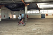Side view of a young Caucasian man with a beard wearing a baseball cap, shorts and a hoodie sitting backwards on the handlebars of a BMX bike while practicing tricks in an abandoned warehouse — Stock Photo