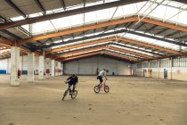 Vista frontal de dos jóvenes caucásicos mirando en direcciones opuestas montando bicicletas BMX mientras practican trucos en un almacén abandonado - foto de stock