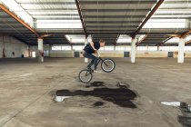 Vue latérale d'un jeune homme caucasien en vélo BMX et sautant du sol, tout en pratiquant des tours dans un entrepôt abandonné — Photo de stock