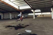 Vista lateral de um jovem caucasiano andando de bicicleta BMX, pulando do chão e girando o guidão, enquanto pratica truques em um armazém abandonado — Fotografia de Stock