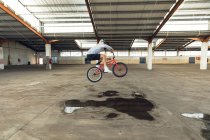 Vista laterale di un giovane caucasico in bicicletta BMX che salta da terra, mentre pratica trucchi in un magazzino abbandonato — Foto stock