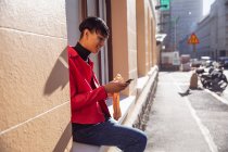 Vista lateral de un joven transexual de raza mixta de moda en la calle, usando un teléfono inteligente y comiendo un sándwich, sentado en un alféizar de la ventana - foto de stock