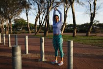 Vorderansicht einer jungen kaukasischen Frau in Sportkleidung, die ihre Arme hebt und sich dehnt, während sie in einem Park trainiert — Stockfoto