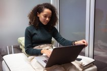Передній вид поруч з молодою змішаною расою жінка сидить у вітальні, працює на ноутбуку комп'ютера в офісі творчого бізнесу — стокове фото