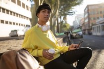Vista frontale di un giovane transgender di razza mista alla moda adulto in strada, con uno smartphone e una tazza di caffè con una bicicletta sullo sfondo — Foto stock