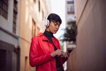 Vista lateral de um jovem elegante mestiço transexual adulto na rua, usando um smartphone com fones de ouvido em — Fotografia de Stock