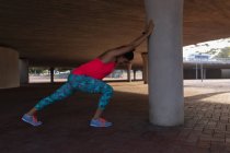 Vista laterale di una giovane donna caucasica che indossa vestiti sportivi spingendo contro un palo sotto un ponte e allungandosi durante un allenamento in un parco — Foto stock