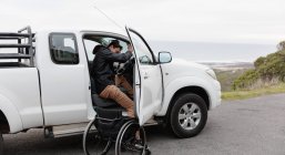 Вид збоку молодого Кавказького чоловіка вихід з інвалідного крісла і в свій автомобіль на автостоянці біля моря — стокове фото