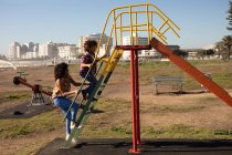 Vista lateral de una joven mestiza y su hijo preadolescente disfrutando del tiempo juntos jugando en un parque infantil junto al mar, subiendo a un tobogán en un día soleado - foto de stock
