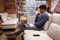 Seitenansicht eines jungen kaukasischen Mannes, der mit einem Laptop auf einem Sofa im Loungebereich eines kreativen Büros sitzt, während seine Kollegen im Hintergrund an Schreibtischen arbeiten. — Stockfoto