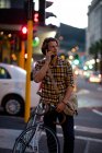 Vue de face gros plan d'un jeune homme caucasien tenant un vélo et parlant sur un téléphone debout dans une rue animée de la ville dans la soirée — Photo de stock