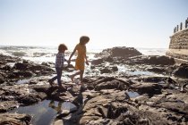 Seitenansicht einer jungen Mischlingsfrau und ihres Sohnes, die die gemeinsame Zeit am Meer genießen, Händchen halten und an einem sonnigen Tag spazieren gehen — Stockfoto
