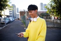 Vue latérale d'un jeune transgenre mixte à la mode adulte dans la rue, utilisant un smartphone et souriant — Photo de stock