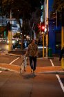Vista frontal de um jovem caucasiano andando na rua com uma bicicleta à noite — Fotografia de Stock