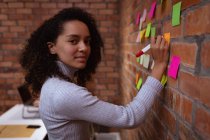 Portrait close up of a young mixed race woman working in the office of a creative business writing on colored sticky notes stuck to an exposed brick wall, turning and looking to camera — Stock Photo