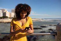 Вид спереду крупним планом молода змішана жінка, використовуючи смартфон у сонячний день біля моря — стокове фото