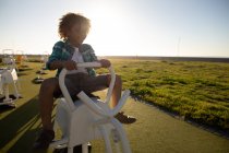Вид спереди на мальчика смешанной расы на детской площадке у моря, сидящего на скакалке в солнечный день — стоковое фото