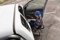 Vista lateral de un joven caucásico en silla de ruedas en la puerta abierta de su camioneta, estacionado en un aparcamiento - foto de stock