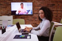 Seitenansicht einer jungen Frau mit gemischter Rasse, die im Büro eines kreativen Unternehmens an einem Laptop arbeitet, sich umdreht und in die Kamera blickt. Ein männlicher Kollege ist auf einem wandmontierten Bildschirm zu sehen, der per Videolink kommuniziert — Stockfoto
