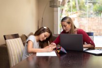 Vista frontale di una giovane donna caucasica che utilizza un computer portatile e la aiuta tra figlia con i compiti nella loro sala da pranzo — Foto stock