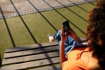 Blick über die Schulter einer jungen Mischlingshündin, die an einem sonnigen Tag auf einer Bank sitzt und ein Smartphone benutzt — Stockfoto