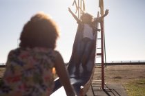 Vista frontal de una joven mestiza y su hijo preadolescente disfrutando del tiempo juntos jugando en un parque infantil junto al mar, el niño deslizándose sobre un tobogán con los brazos en el aire en un día soleado - foto de stock