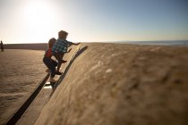 Vista lateral de uma jovem mulher de raça mista e seu filho pré-adolescente desfrutando de tempo juntos junto ao mar, escalando uma parede em um dia ensolarado — Fotografia de Stock