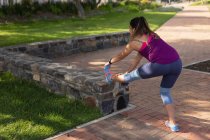 Rear view of a young Caucasian woman wearing sports clothes touching her toes with her leg up on a low wall during a workout in a park — Stock Photo